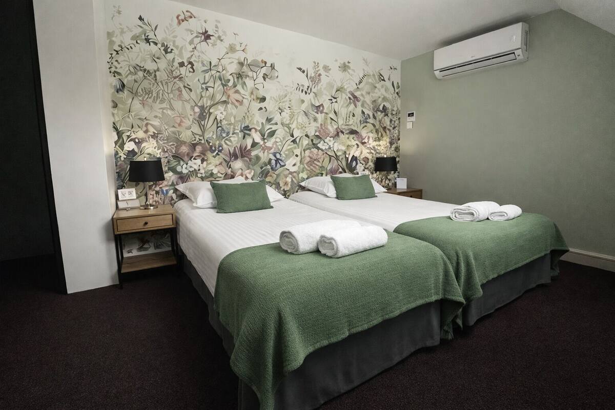 A hotel room with two beds, green bedspreads, a floral wallpaper, a nightstand with a lamp, and an air conditioning unit.