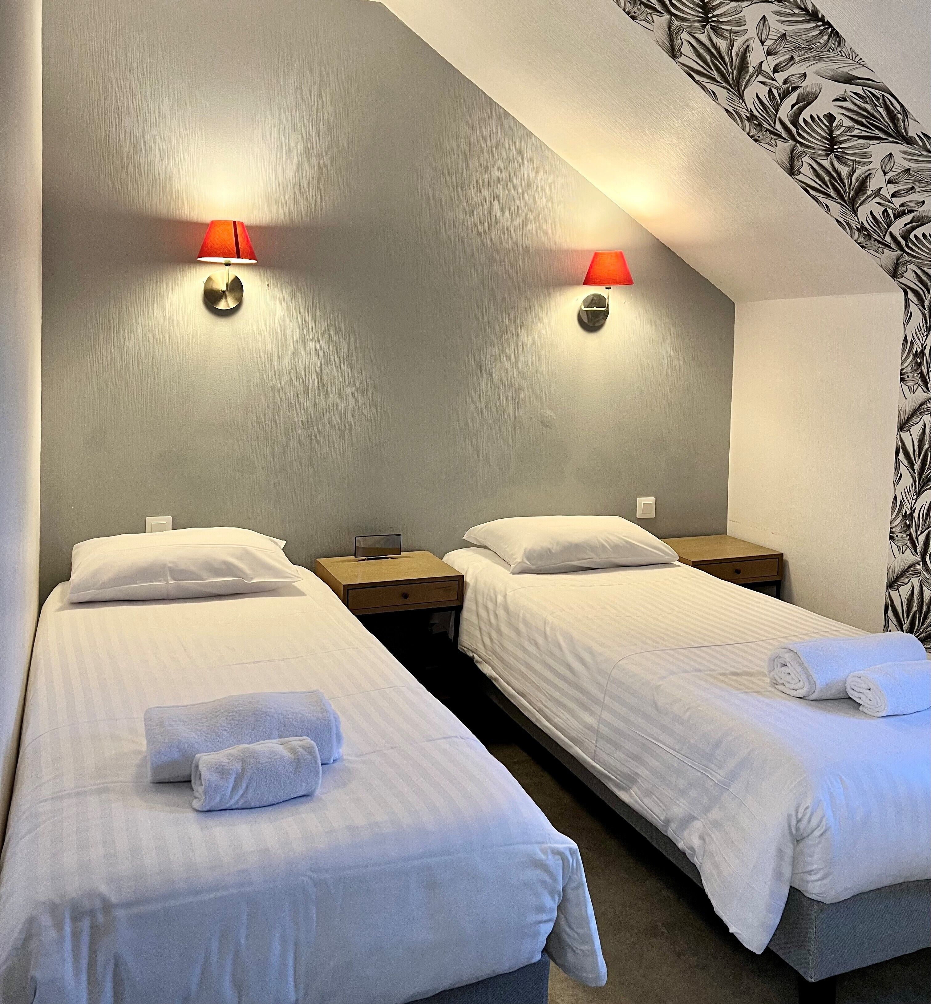 A hotel room with two single beds, each with a pillow and a rolled towel. There are two wall-mounted lamps with red shades.