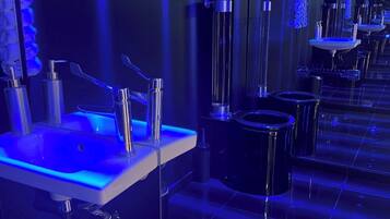 A modern bathroom with multiple sinks and toilets, illuminated by blue lighting.