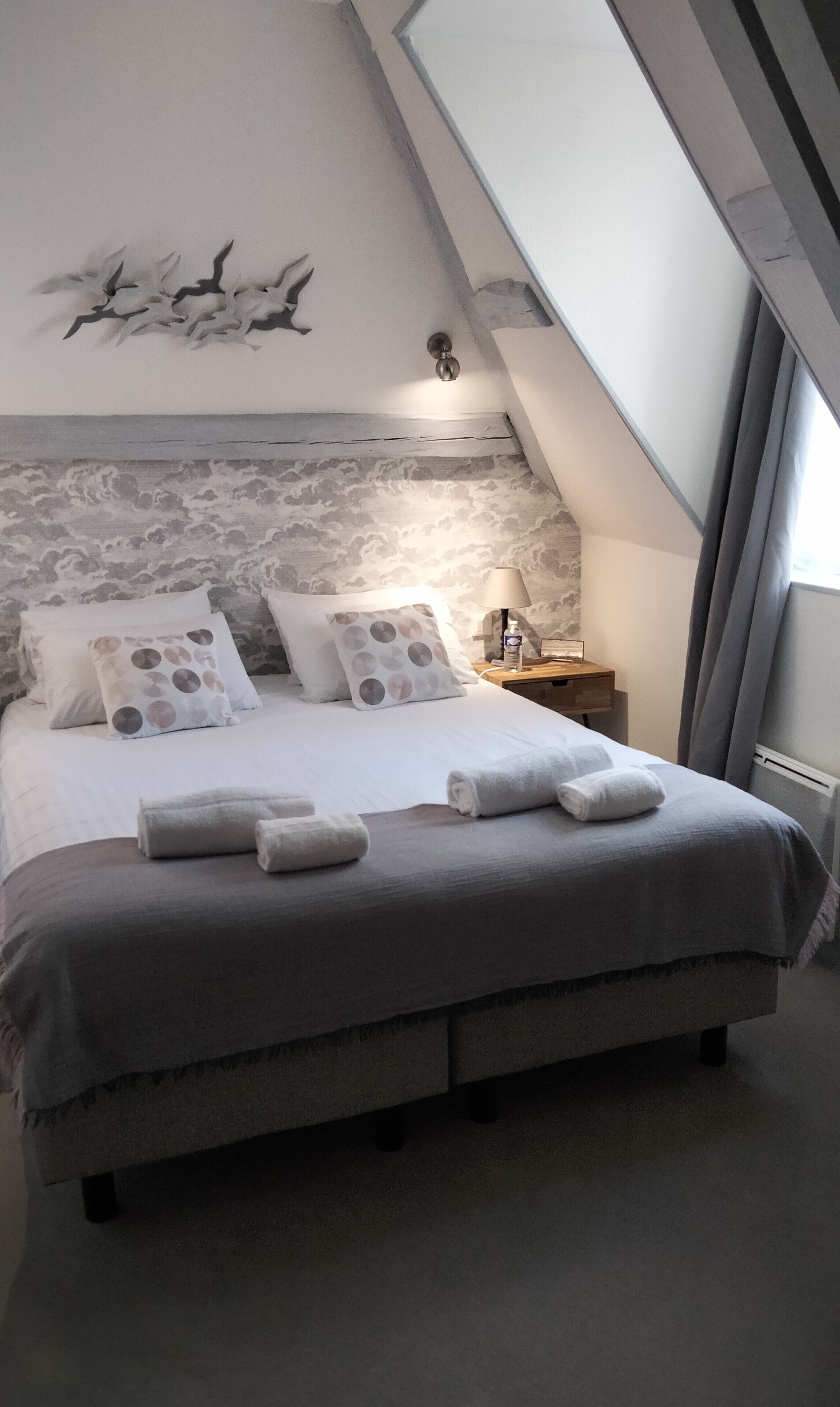 A bedroom with a bed, pillows, a nightstand, a lamp, and a wall with a bird mural.