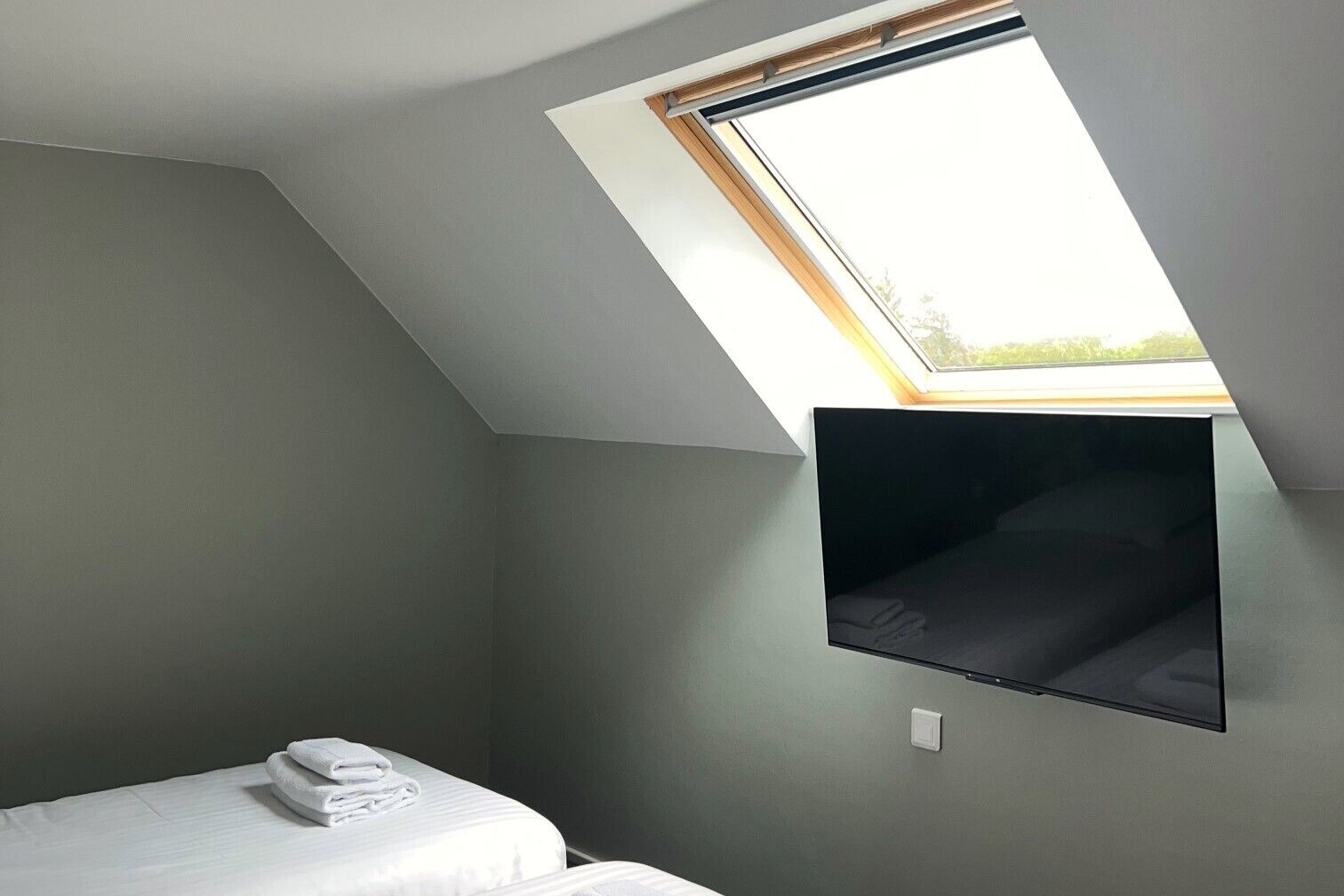 A room with a bed, a flat-screen TV mounted on the wall, and a skylight.