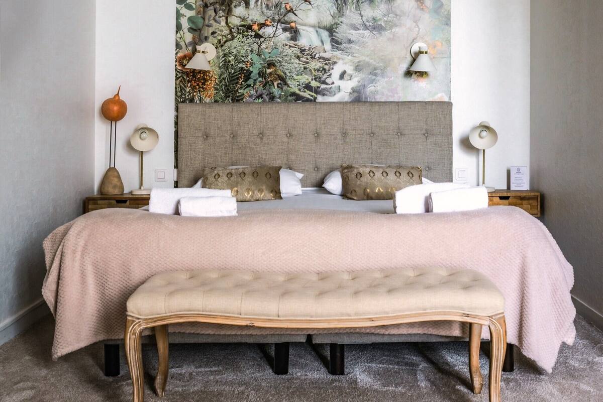 A bedroom with a large bed, two nightstands, a bench, and a wall-mounted lamp.
