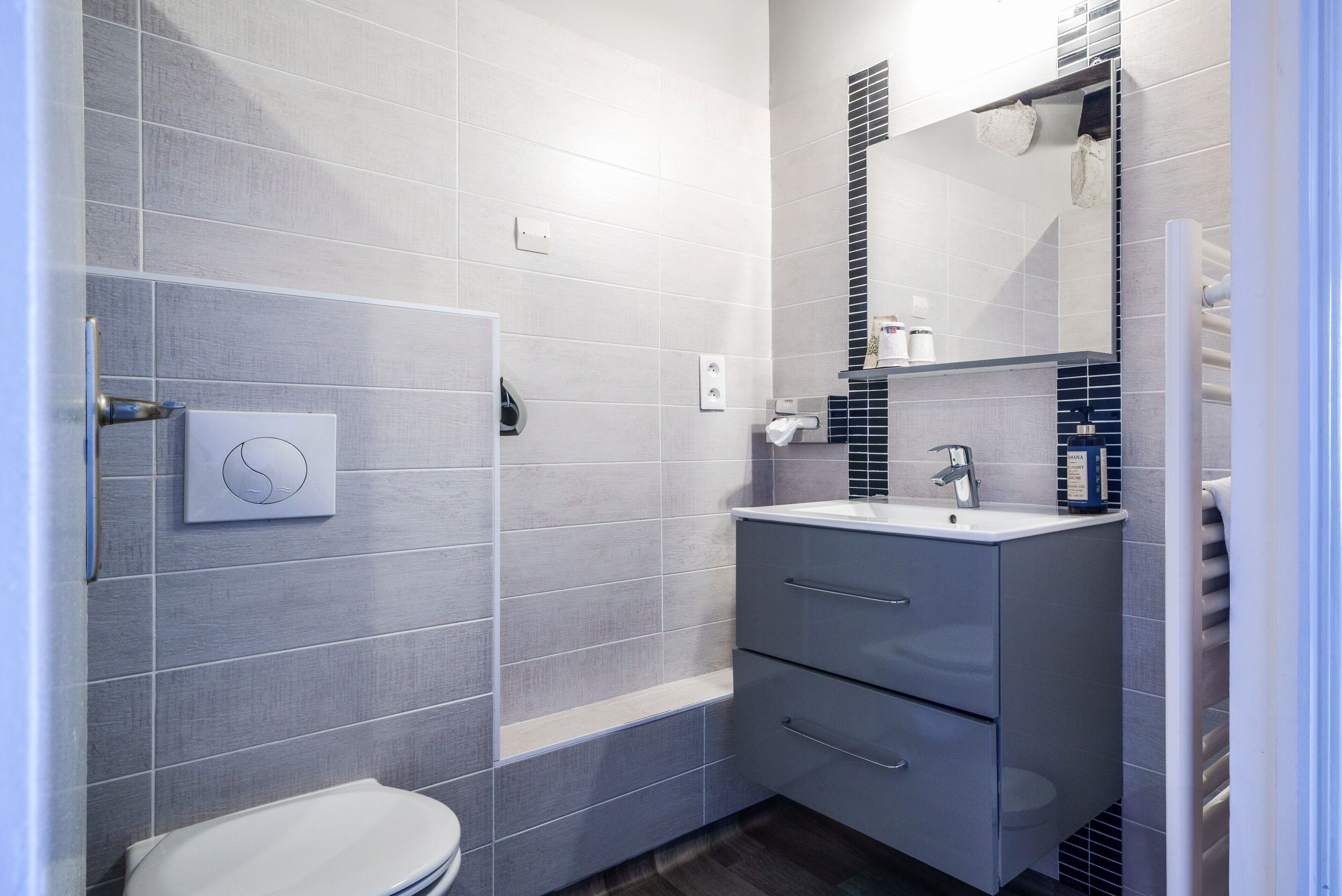 A modern bathroom with grey tiles, a toilet, a sink with a mirror, and a towel dispenser.