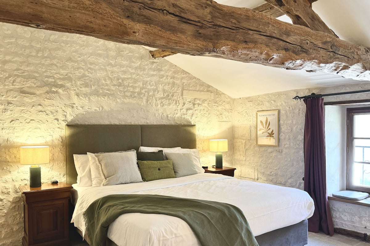 A bedroom with a stone wall, wooden beams, a bed with white bedding, two bedside tables with lamps, and a window with curtains.