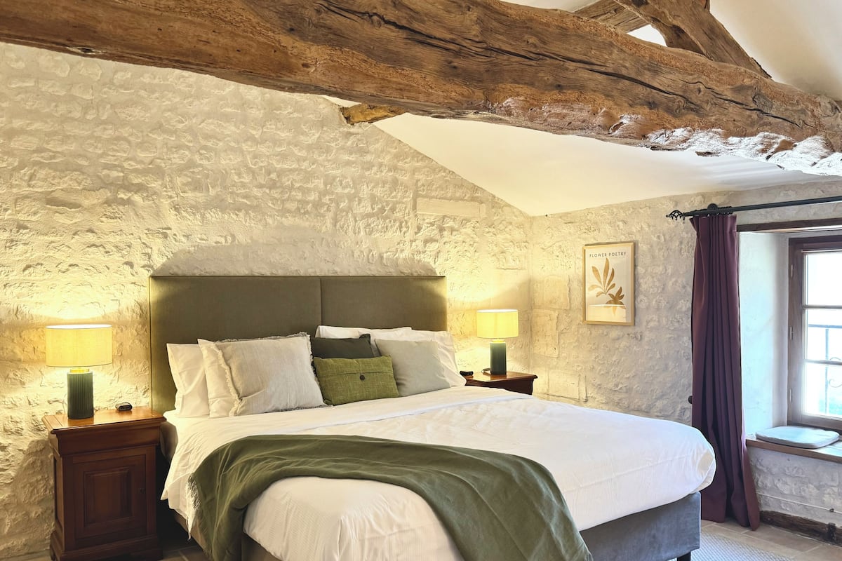 A bedroom with a stone wall, wooden beams, a bed with white bedding, two bedside tables with lamps, and a window with curtains.