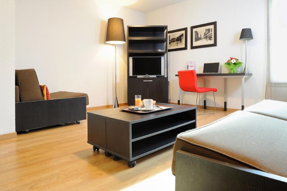 A modern hotel room with a sofa, a desk with a computer, a flat-screen TV, and a coffee table.