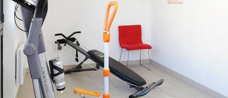 A gym with exercise equipment including a stationary bike, elliptical, and treadmill. There are green mats on the floor, a red chair, and artwork on the walls.