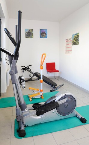 Un gimnasio con equipo de ejercicio, incluyendo una bicicleta estática, elíptica y cinta de correr. Hay colchonetas verdes en el suelo, una silla roja y obras de arte en las paredes.