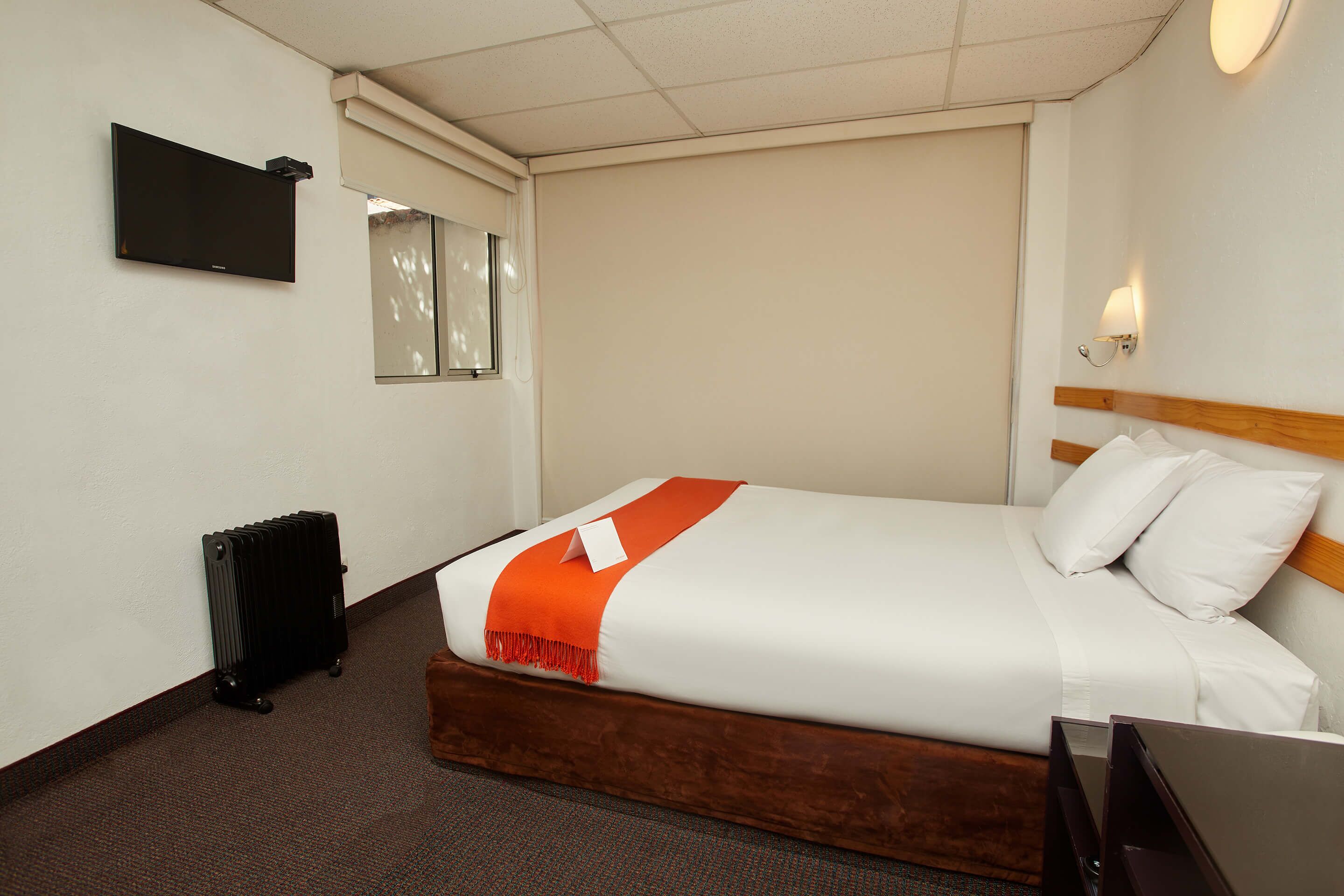 A hotel room with a bed, a television, a radiator, and a wall-mounted light.