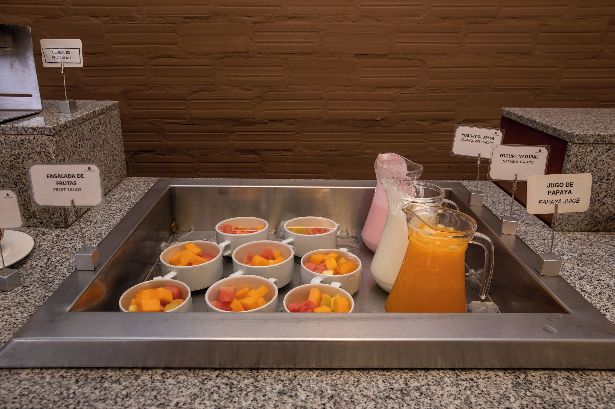 Una estación de bebidas de autoservicio con recipientes etiquetados para cereales, ensalada de frutas, yogur y jugo de papaya.