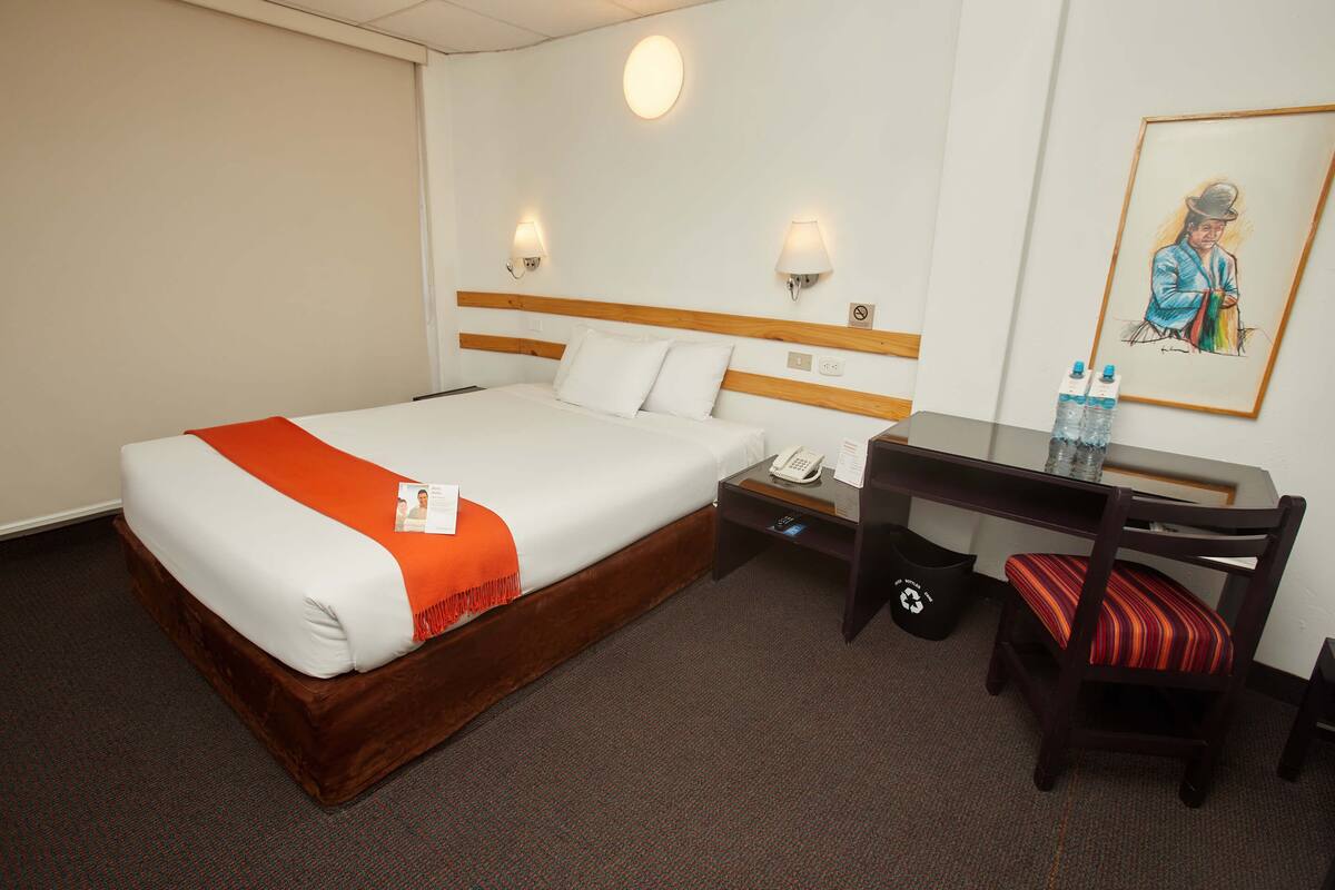 A hotel room with a bed, a desk, a chair, a telephone, and a trash bin.