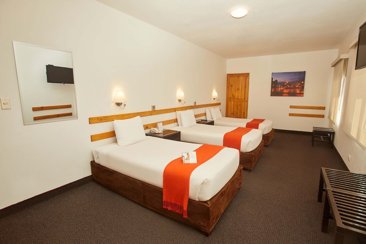 A hotel room with three beds, a wooden headboard, a flat-screen TV, a nightstand, a small table, and a window with a cityscape view.