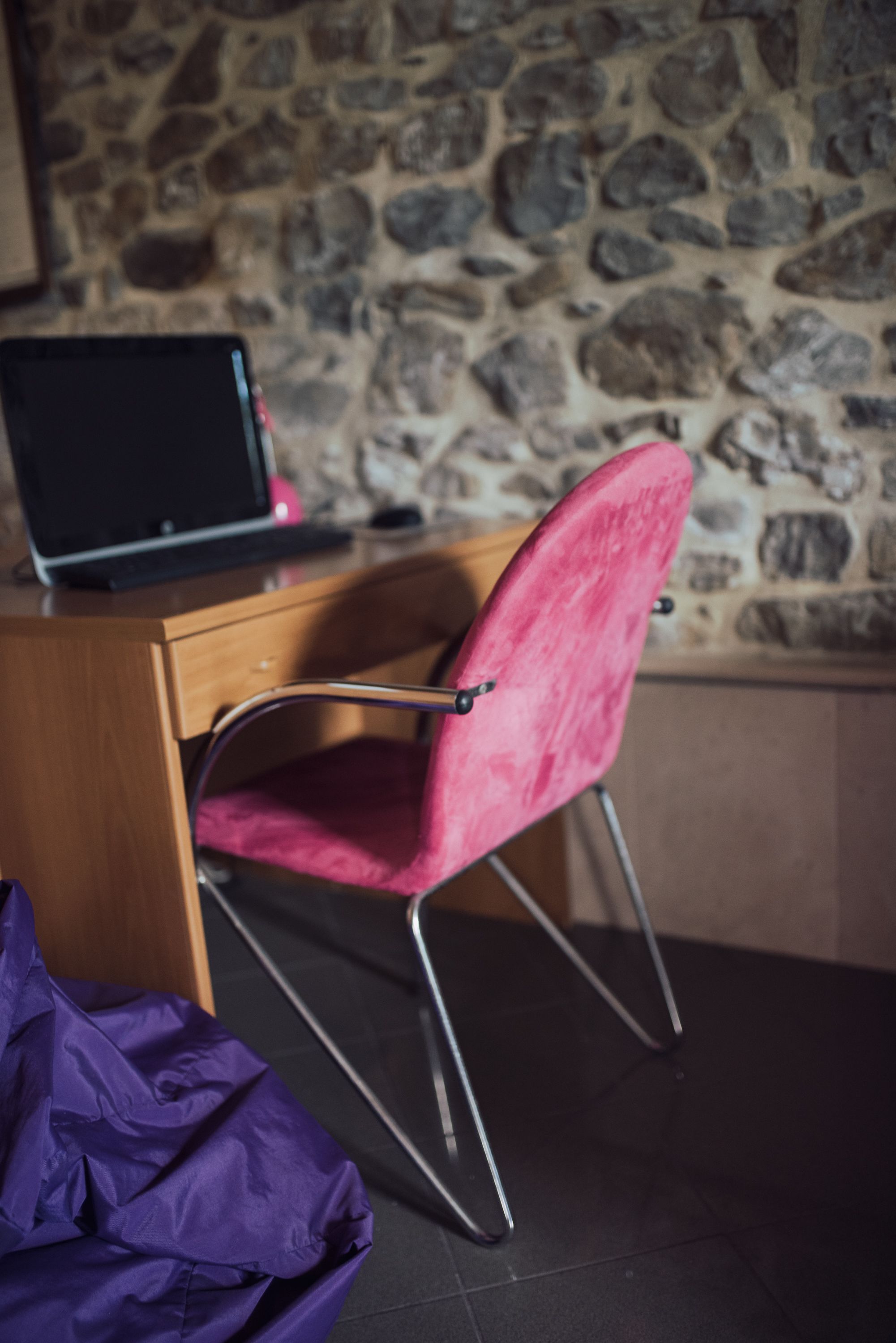 Une pièce avec un mur en pierre, un bureau en bois, une chaise rose et une couverture violette sur le sol.
