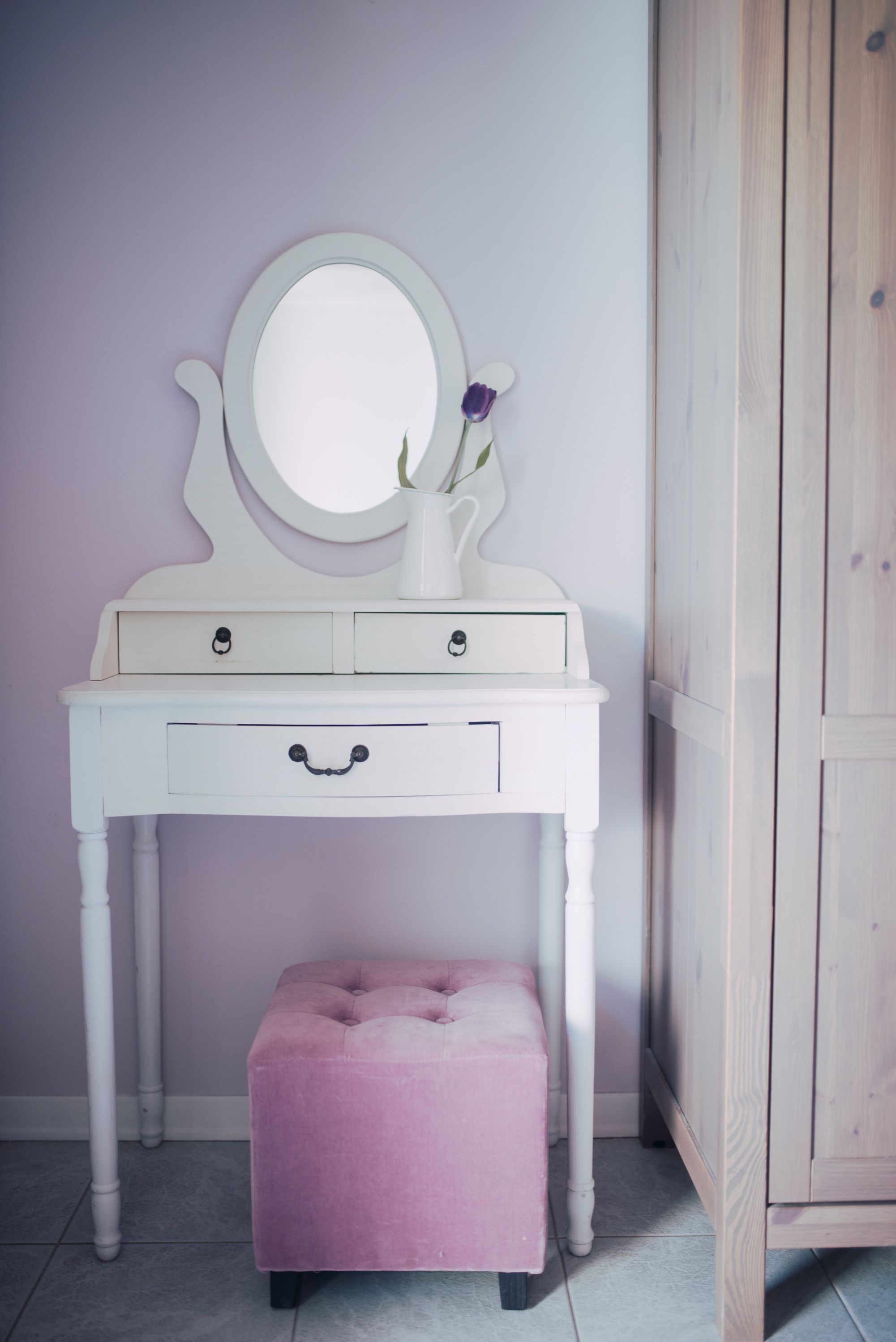 A white vanity with a round mirror, two drawers, and a pink ottoman.