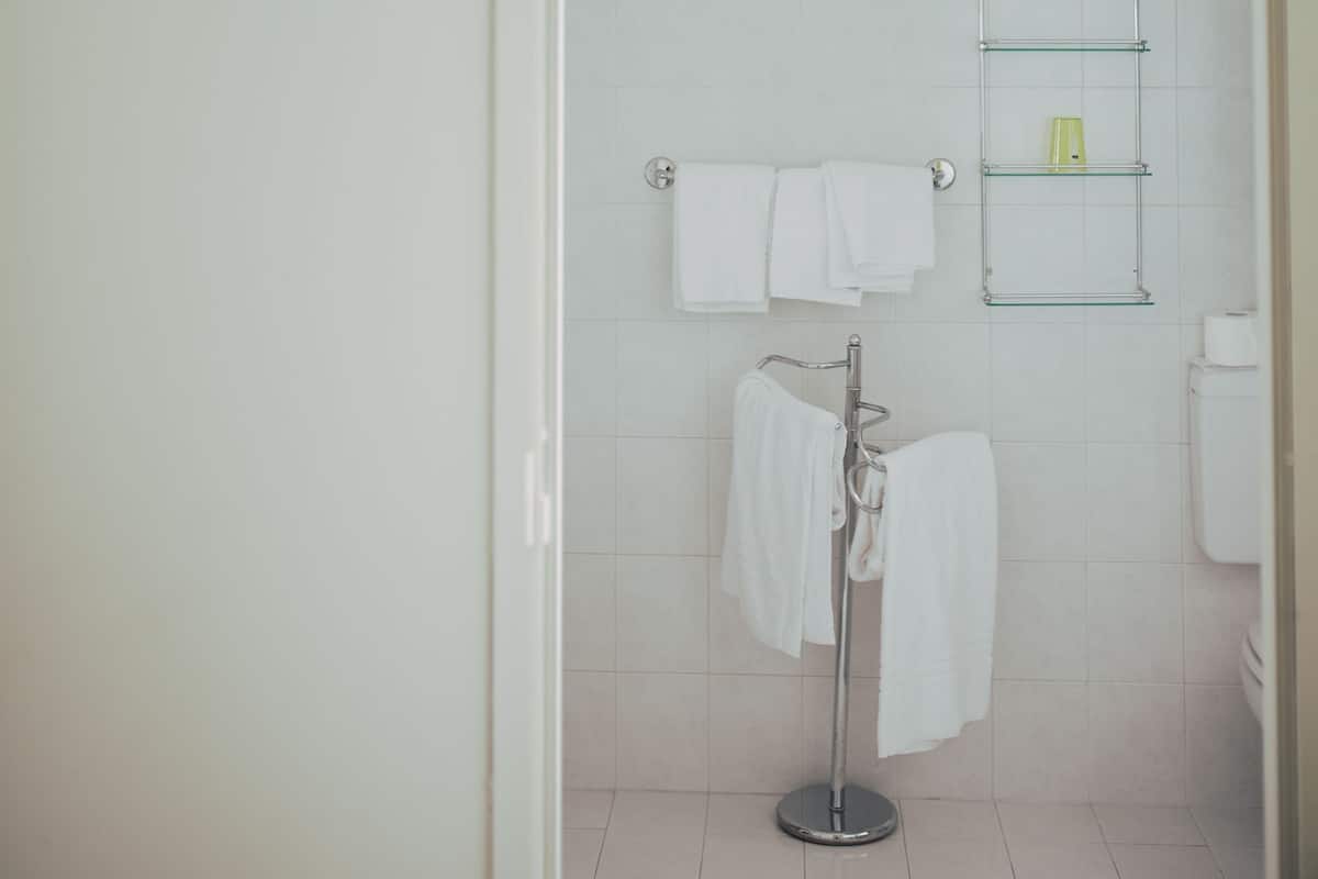 A bathroom with a white tiled wall, a glass shower enclosure, a towel rack with white towels, and a glass shelf with toiletries.