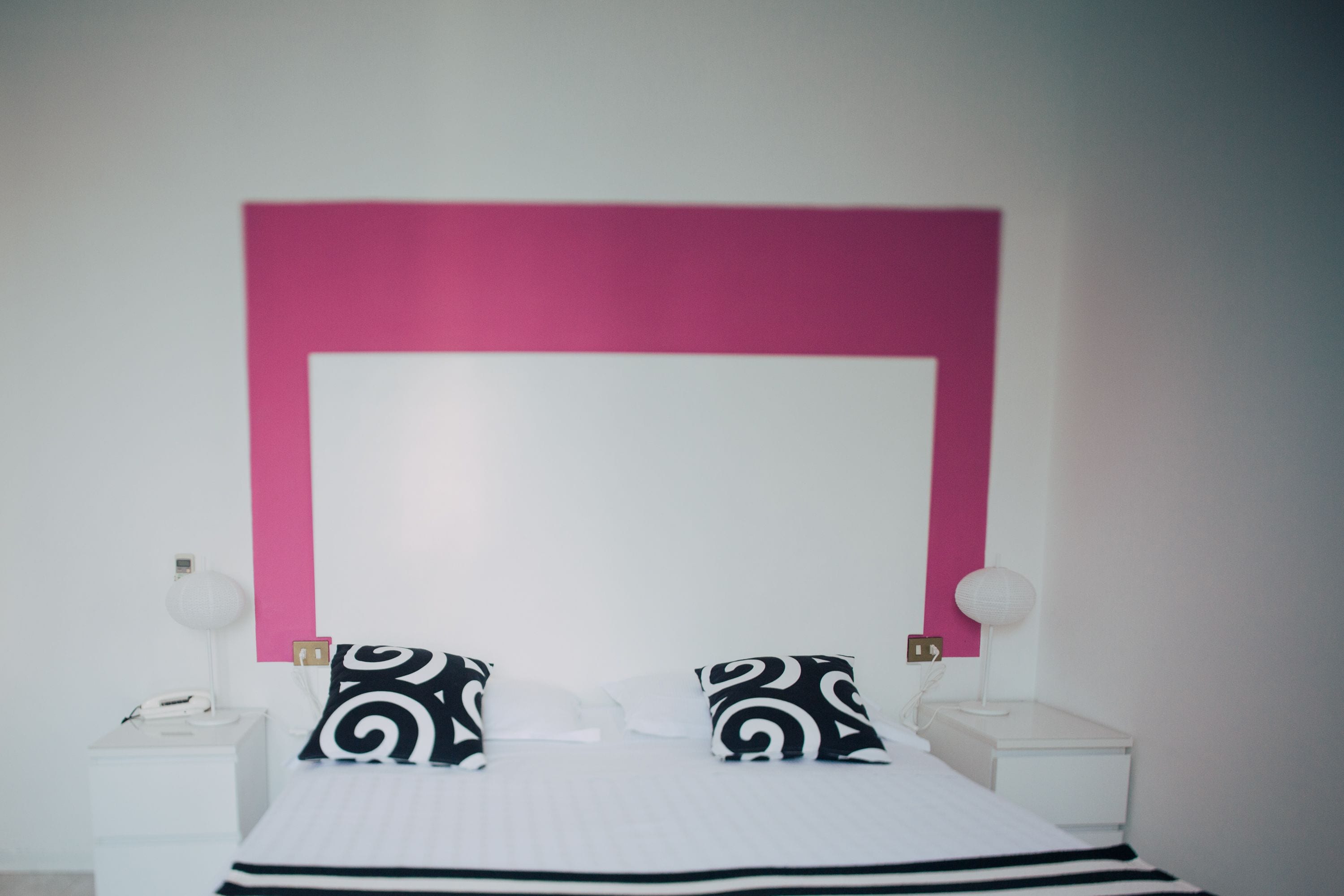 A bed with a white and black striped bedspread, two black and white patterned pillows, a white bedside table with a lamp, and a pink accent wall.