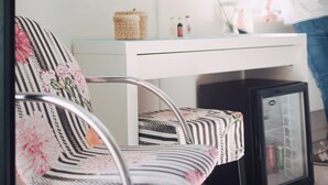 Una habitación moderna con un escritorio blanco, un televisor de pantalla plana montado en la pared, un refrigerador pequeño y una silla con estampado floral.