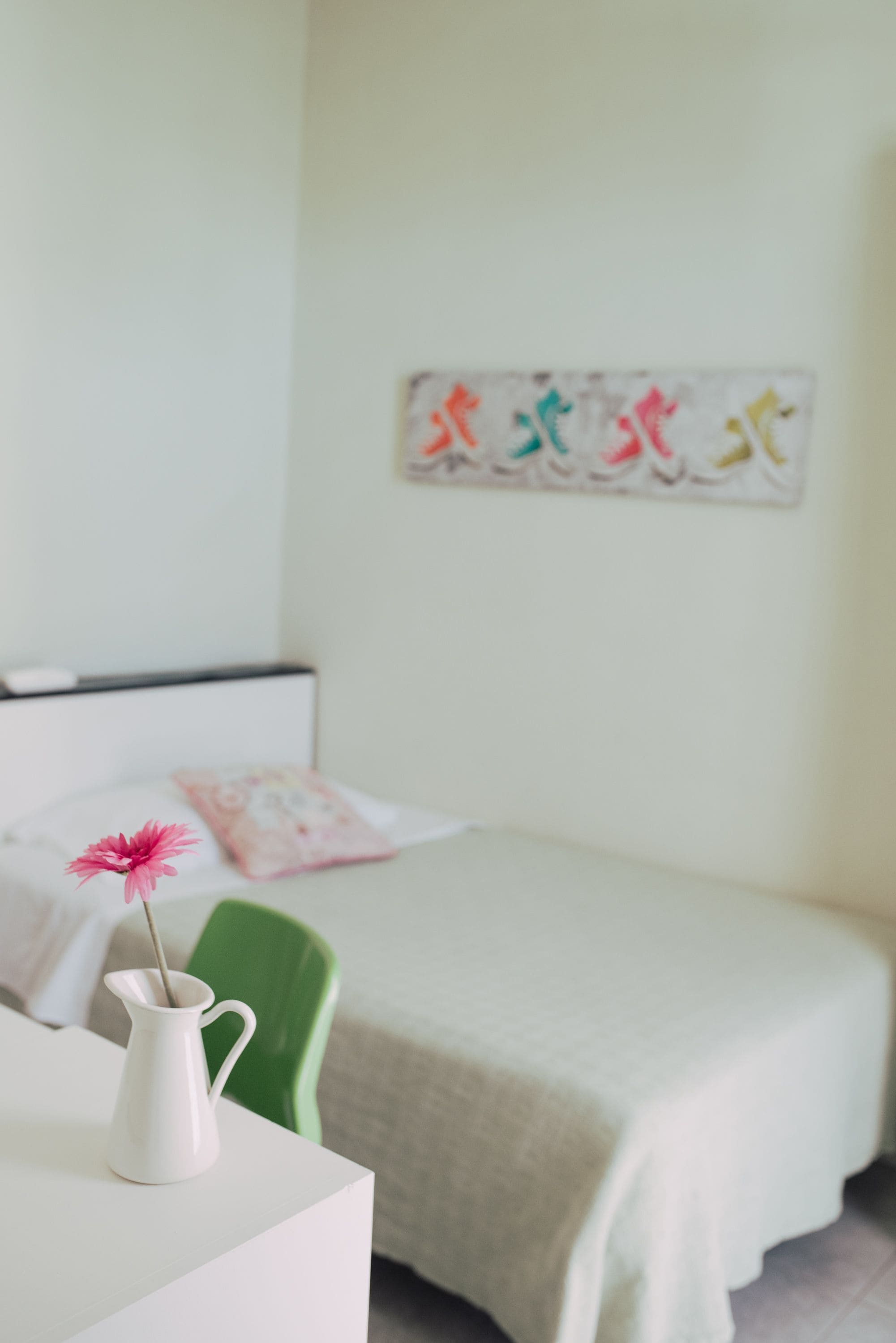 A neatly arranged bedroom with a bed, a green chair, a white bedside table, and a white pitcher with a pink flower.