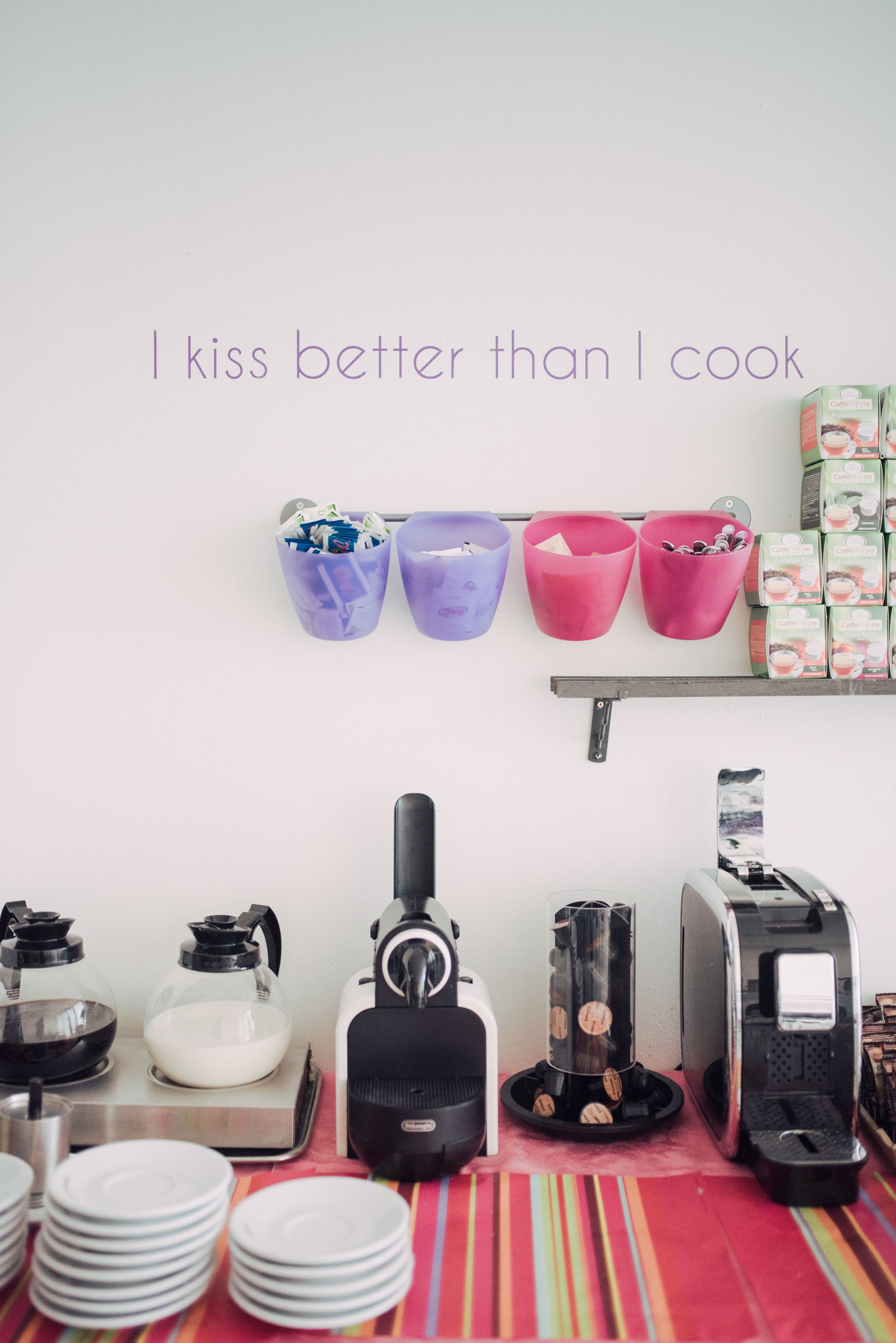 A kitchen area with various appliances and utensils, including a coffee maker, a blender, and a toaster. There are also several containers and a stack of plates on the counter.