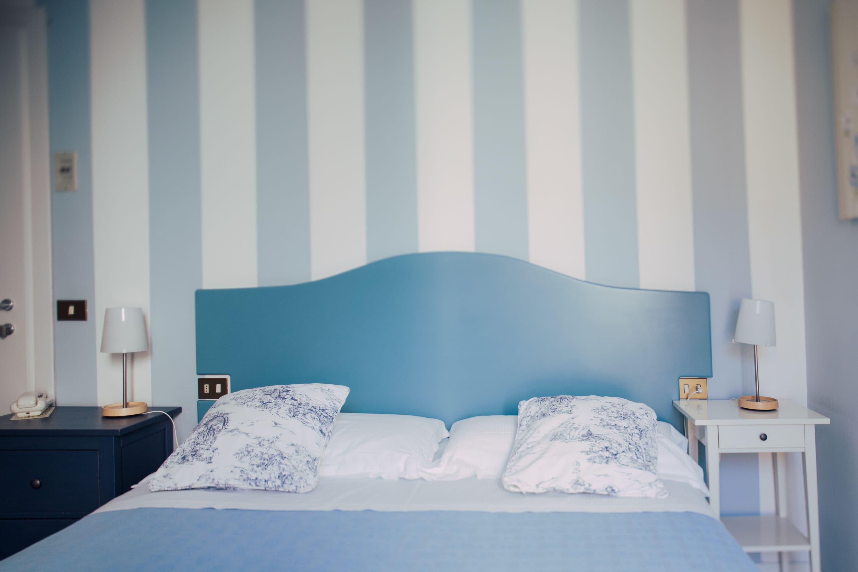 Un lit avec une tête de lit bleue, une literie blanche et des coussins à motifs. De chaque côté du lit, il y a des tables de chevet avec des lampes et un téléphone.