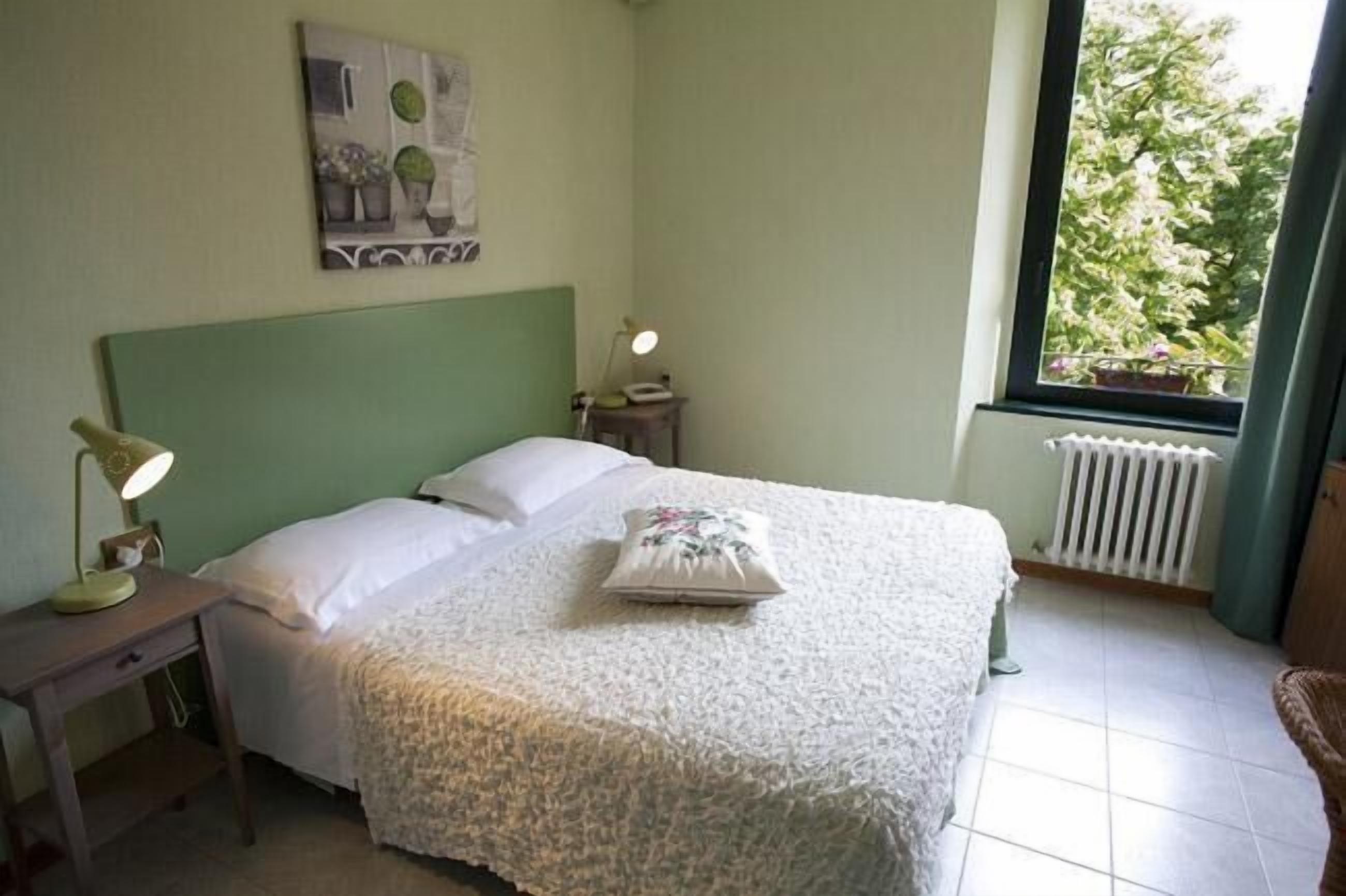 Un dormitorio con cama, mesita de noche, lámpara, radiador y ventana con vista a árboles.
