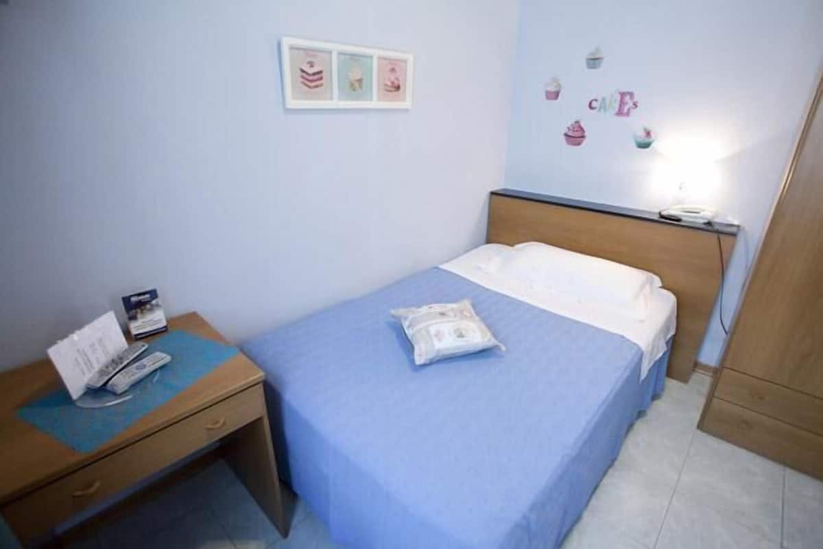 A single bed with a blue bedspread, a bedside table with a remote control, a small framed picture, and a wall with cupcake decorations.