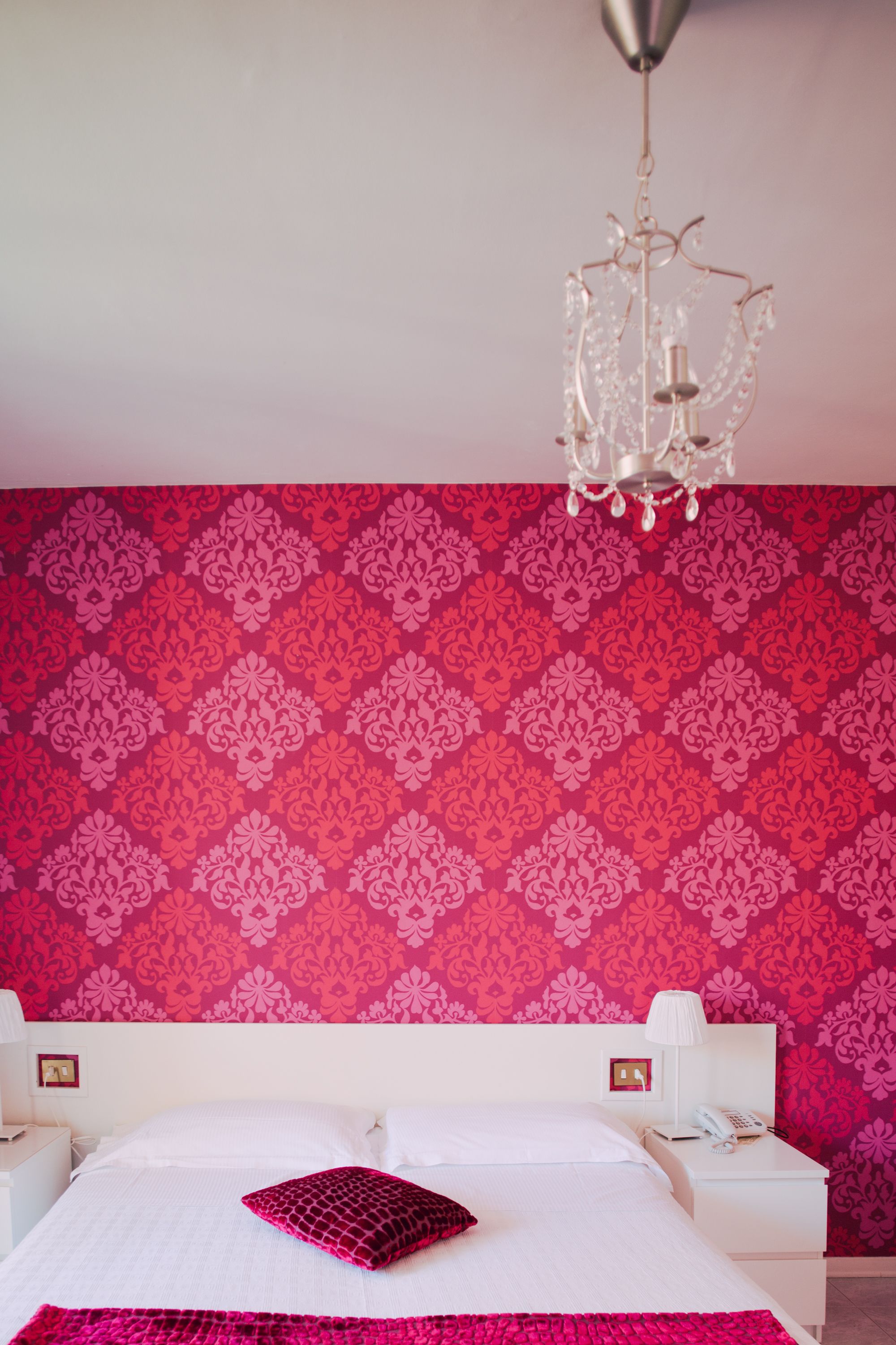 Una camera d'albergo con carta da parati decorata, un lampadario, un letto con lenzuola bianche e un cuscino decorativo rosso.