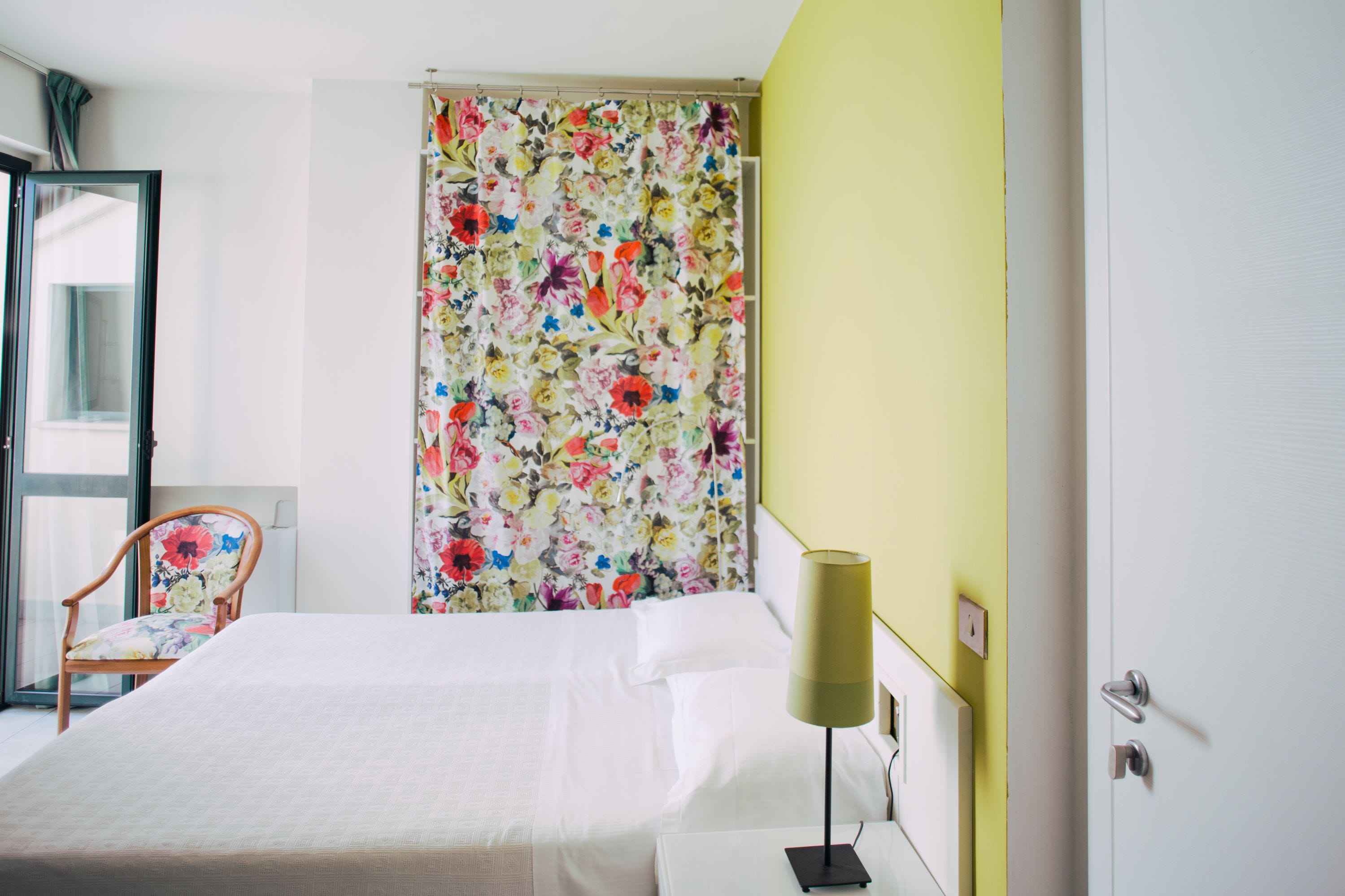 A hotel room with a bed, a chair, a curtain with a floral pattern, and a lamp.