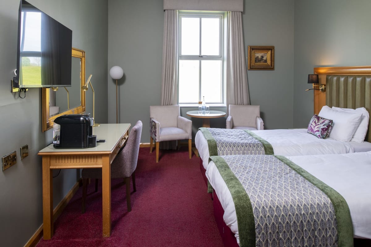 Habitación de hotel con una cama, dos sillas, un escritorio con cafetera, una mesa redonda, una ventana con cortinas y un cuadro en la pared.