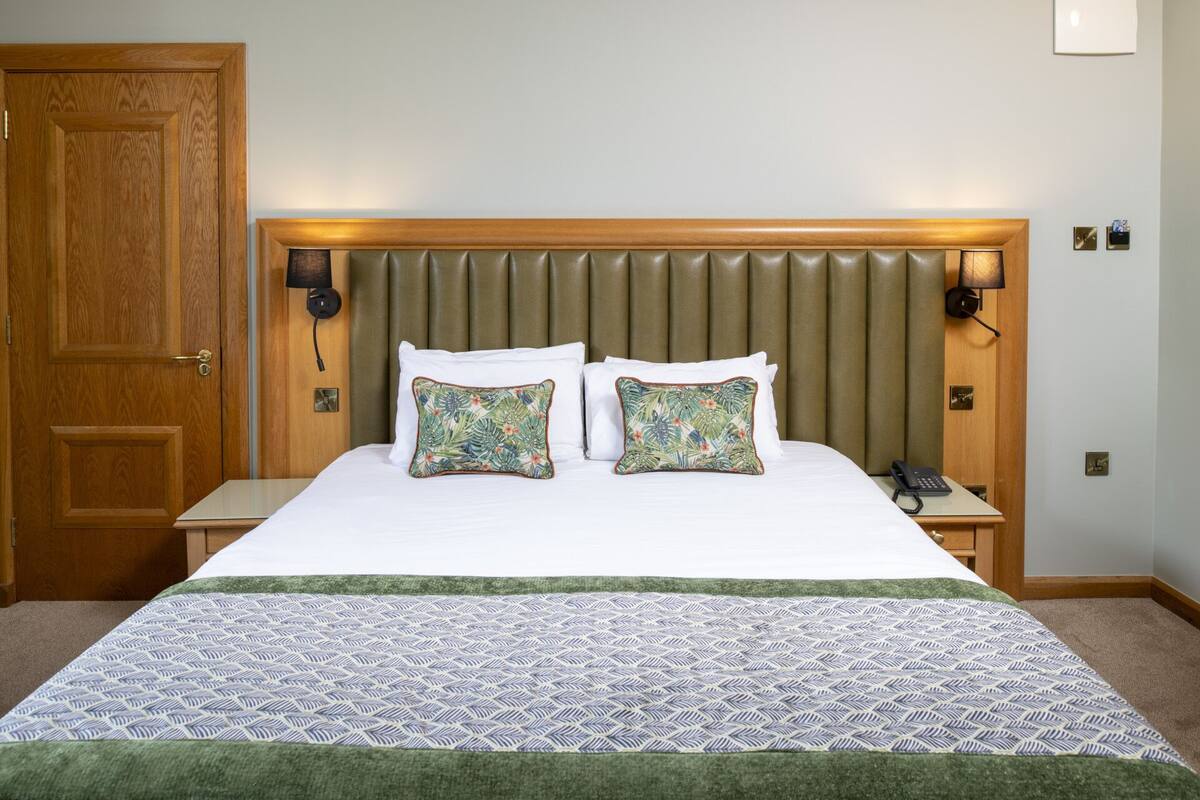 Habitación de hotel con puerta de madera, cama con colcha estampada, dos almohadones decorativos, cabecera con listones verticales y lámparas de noche.