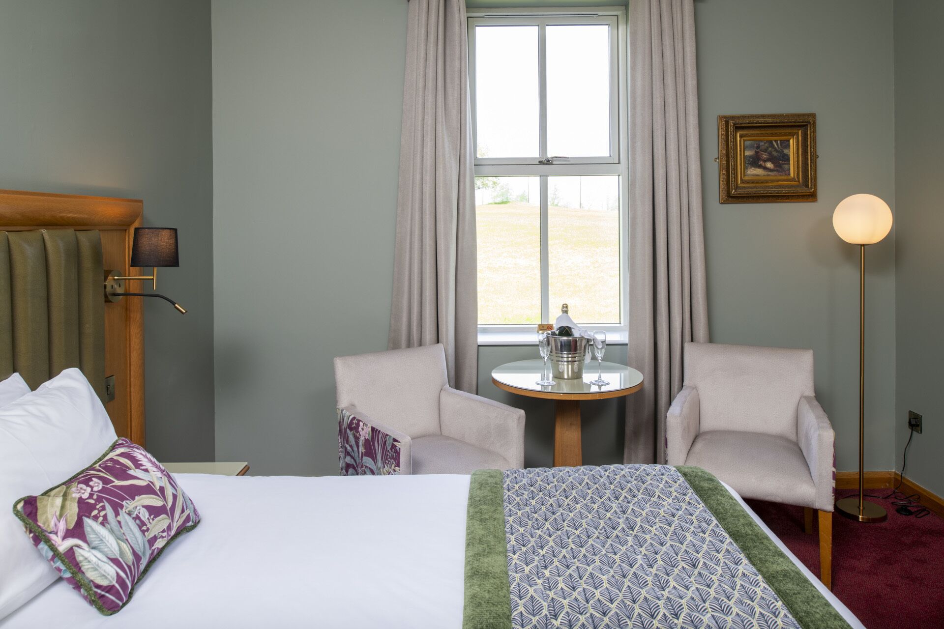 Habitación de hotel con cama, dos sillones, una mesita con una botella y vasos, una lámpara y un cuadro en la pared.