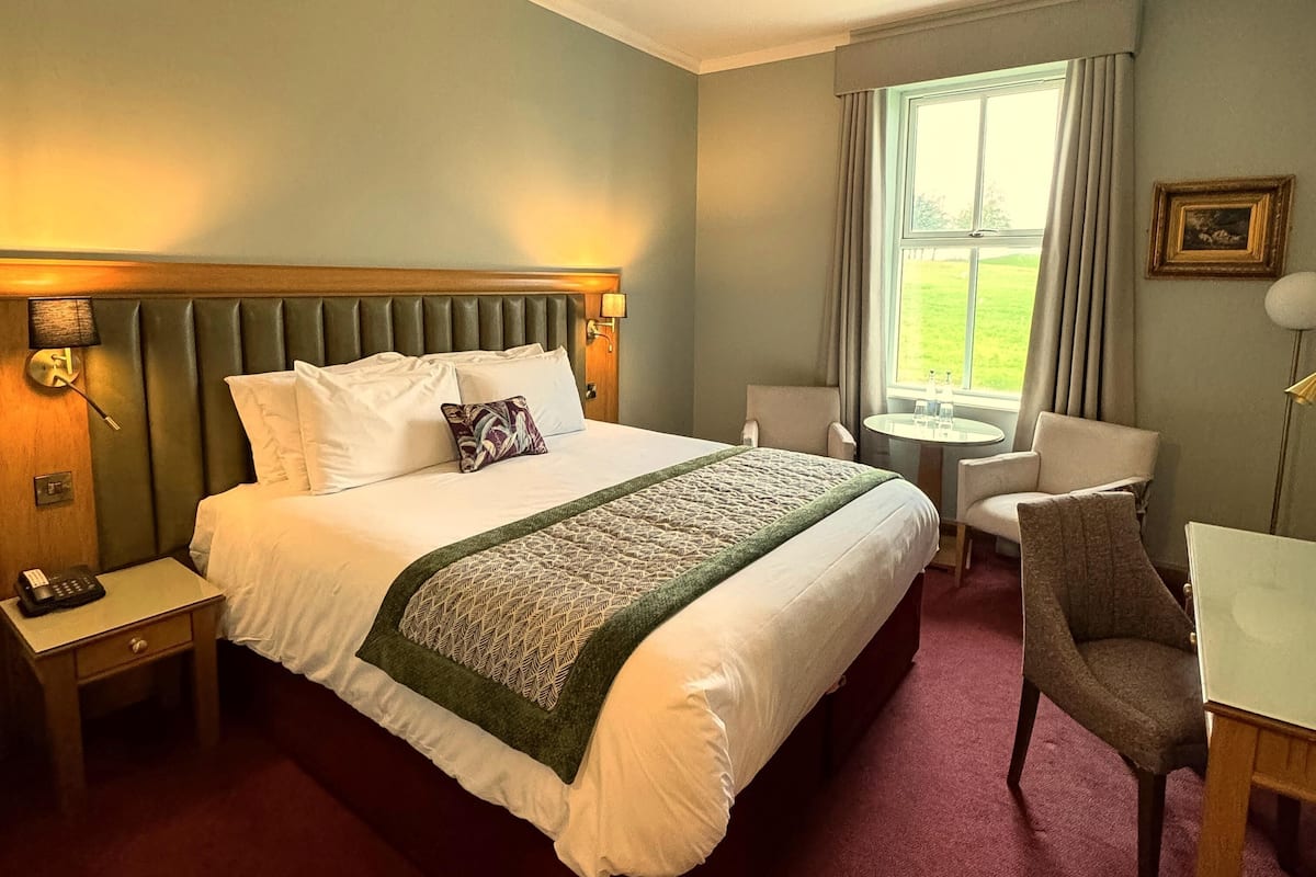 Una habitación de hotel con una cama grande, una mesita de noche con teléfono, una pequeña mesa redonda con dos sillas y una ventana con cortinas.