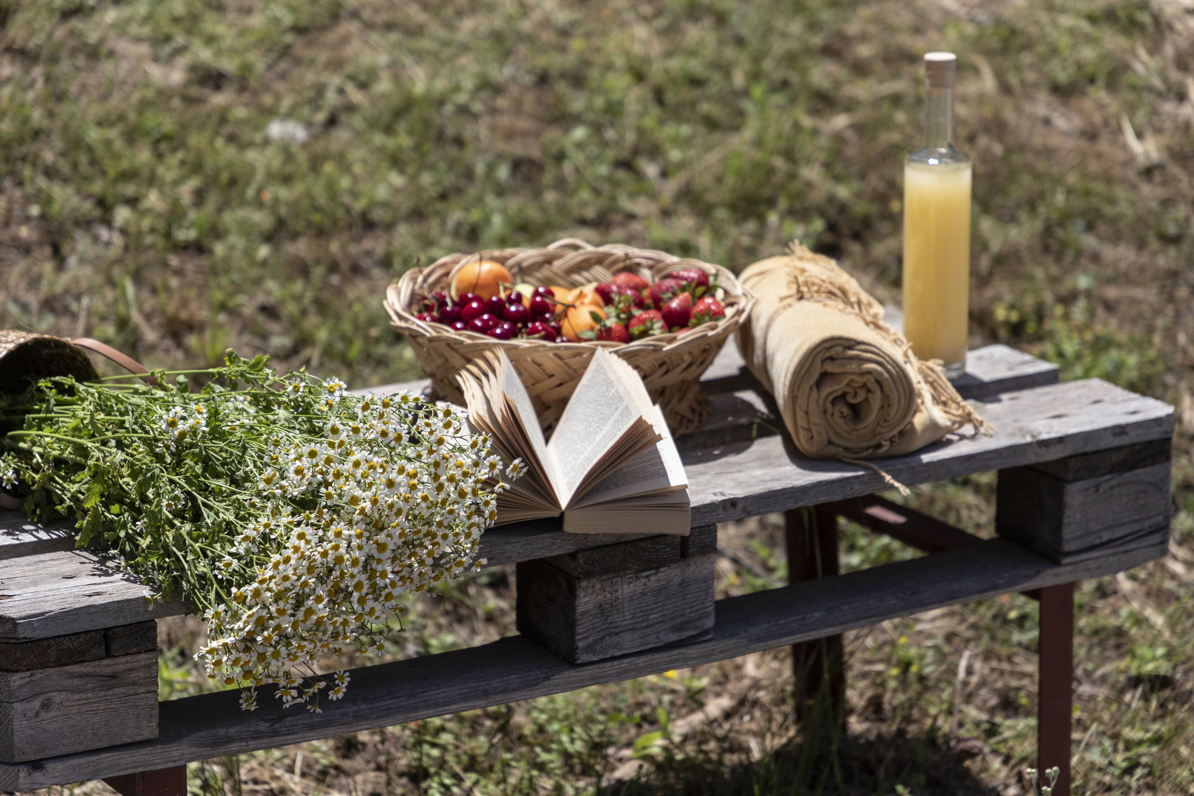 A wooden picnic table with a basket of fruits, a bottle of juice, and a rolled-up cloth.