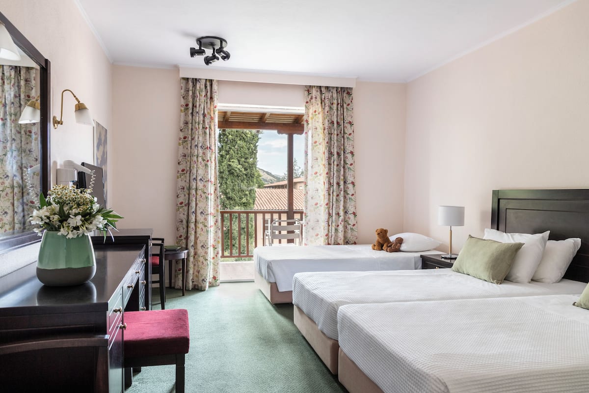 Ein Hotelzimmer mit zwei Betten, einem Schreibtisch mit einer Vase, einem Sessel, einem Nachttisch und einem Balkon mit Vorhängen.