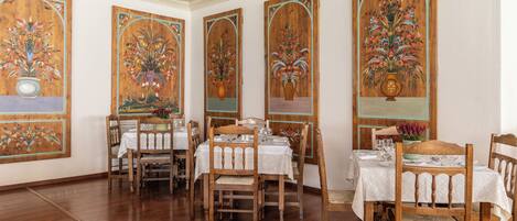 A dining area with wooden tables and chairs, white tablecloths, and floral paintings on the walls.