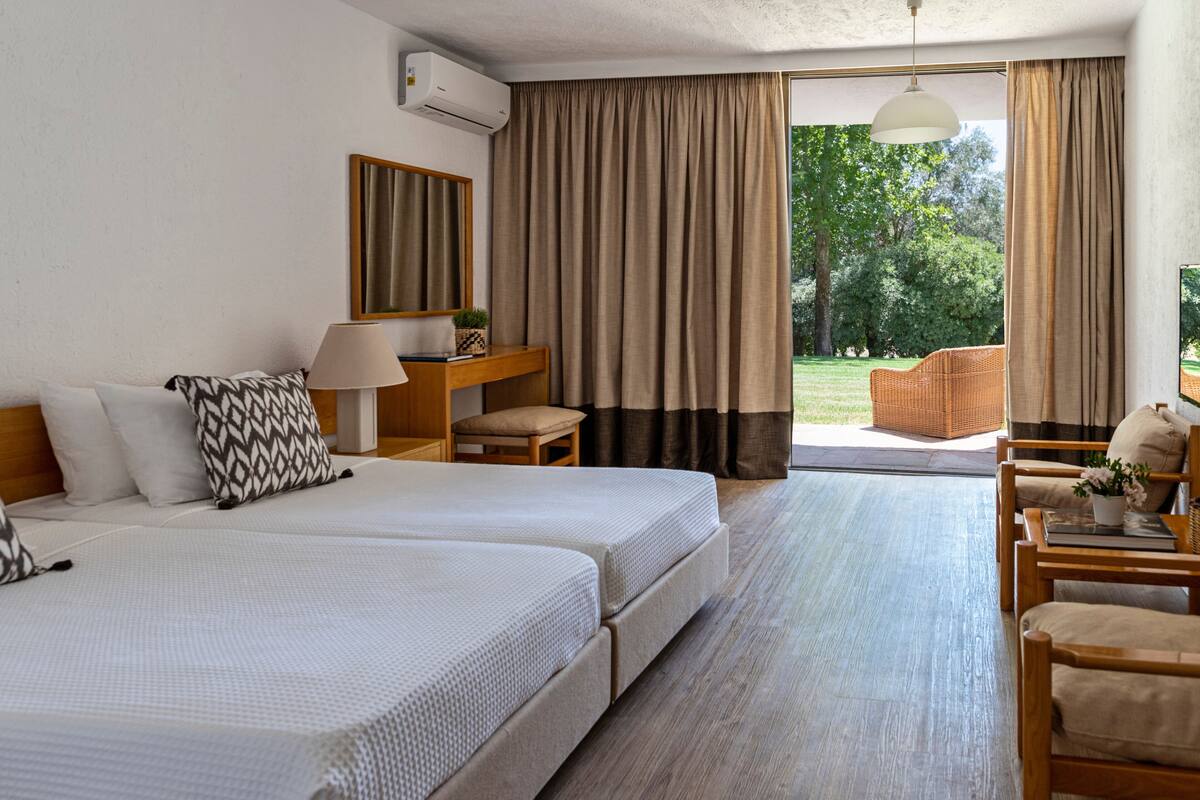 A hotel room with two beds, a desk, a chair, a lamp, and a view of the outdoors.