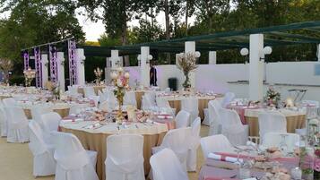Un espacio al aire libre para eventos con mesas redondas dispuestas para comer, rodeado de sillas blancas.