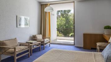 A hotel room with a bed, two chairs, a small table, a balcony with curtains, and a view of trees.