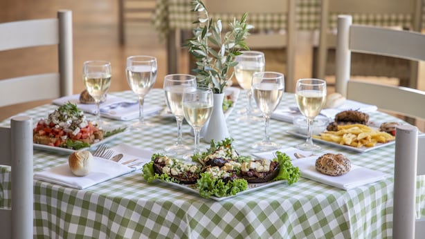 Mesa posta com toalha xadrez, pratos com comida, copos de vinho branco e um arranjo de folhagens.