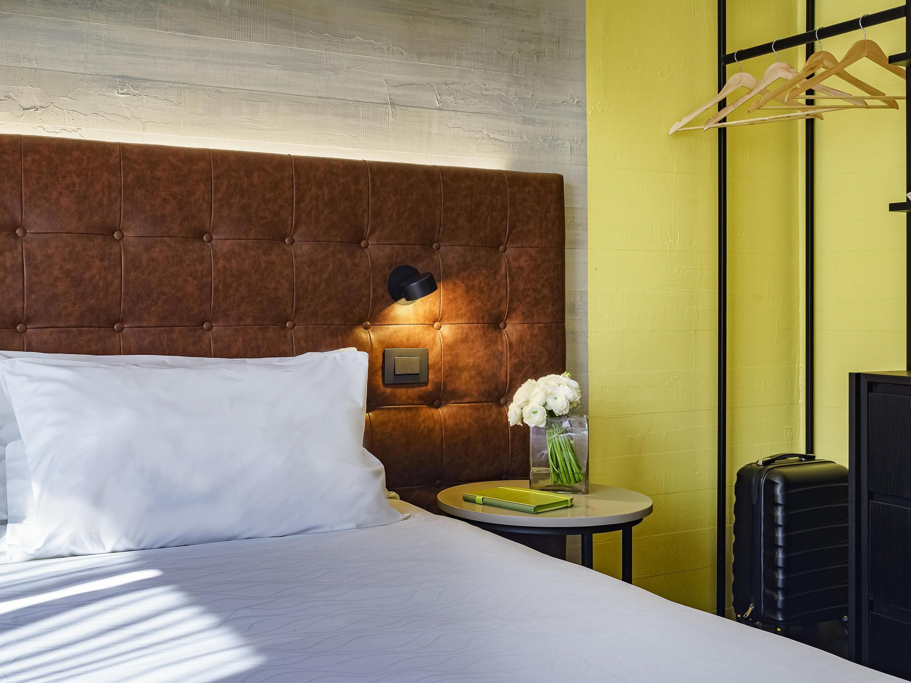 Habitación de hotel con tablero de cabecera capitonado, mesita de noche con un florero, maleta y perchero.