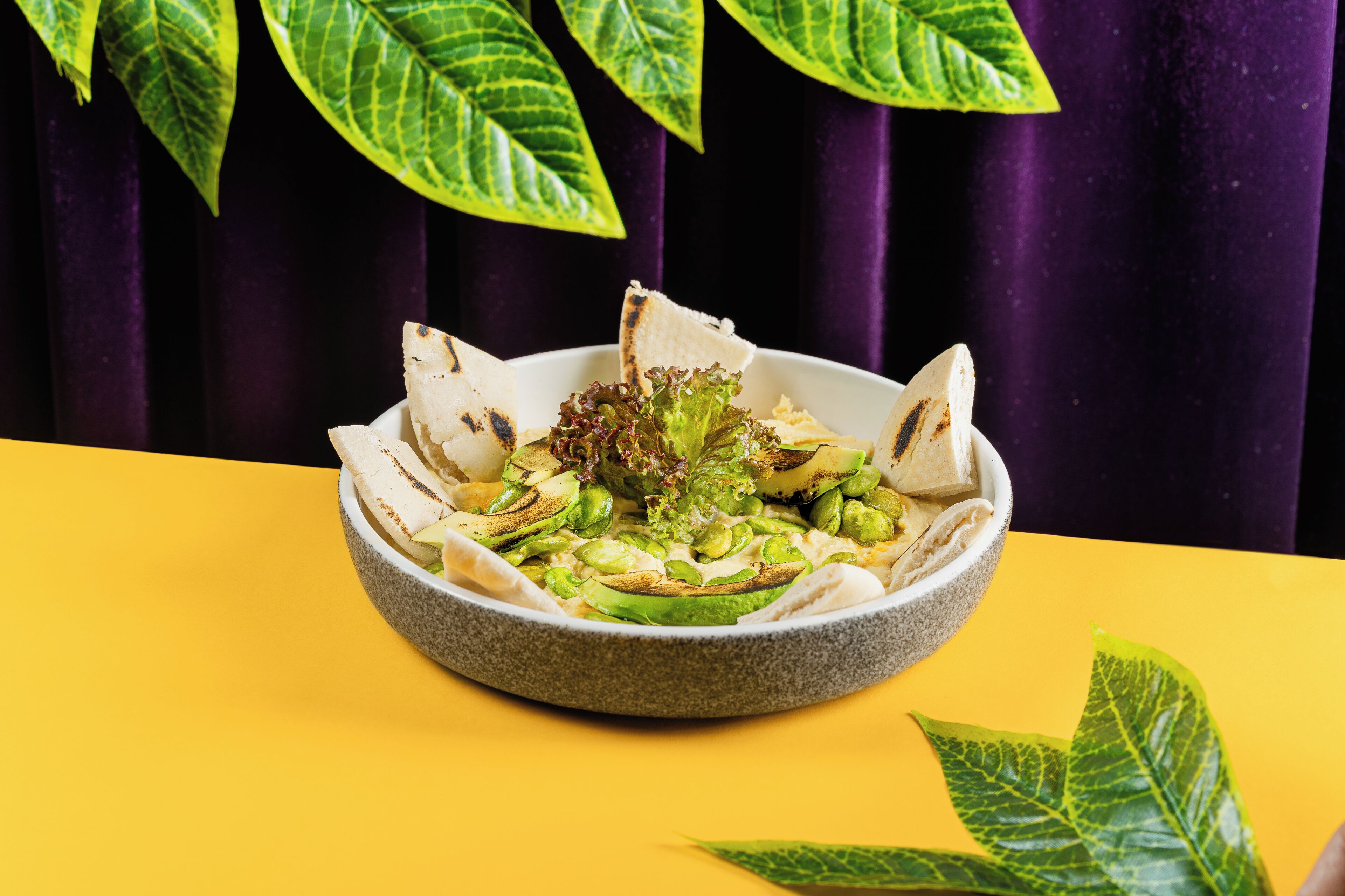 A bowl of salad on a yellow table with a purple curtain in the background.