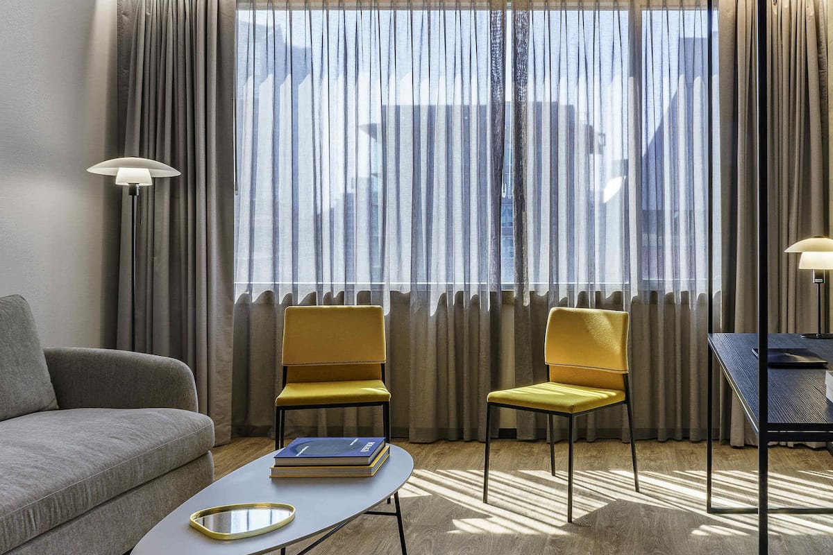 Ein modernes Hotelzimmer mit einer Couch, zwei gelben Sesseln, einem kleinen Tisch mit Büchern und einem Schreibtisch mit Lampe.