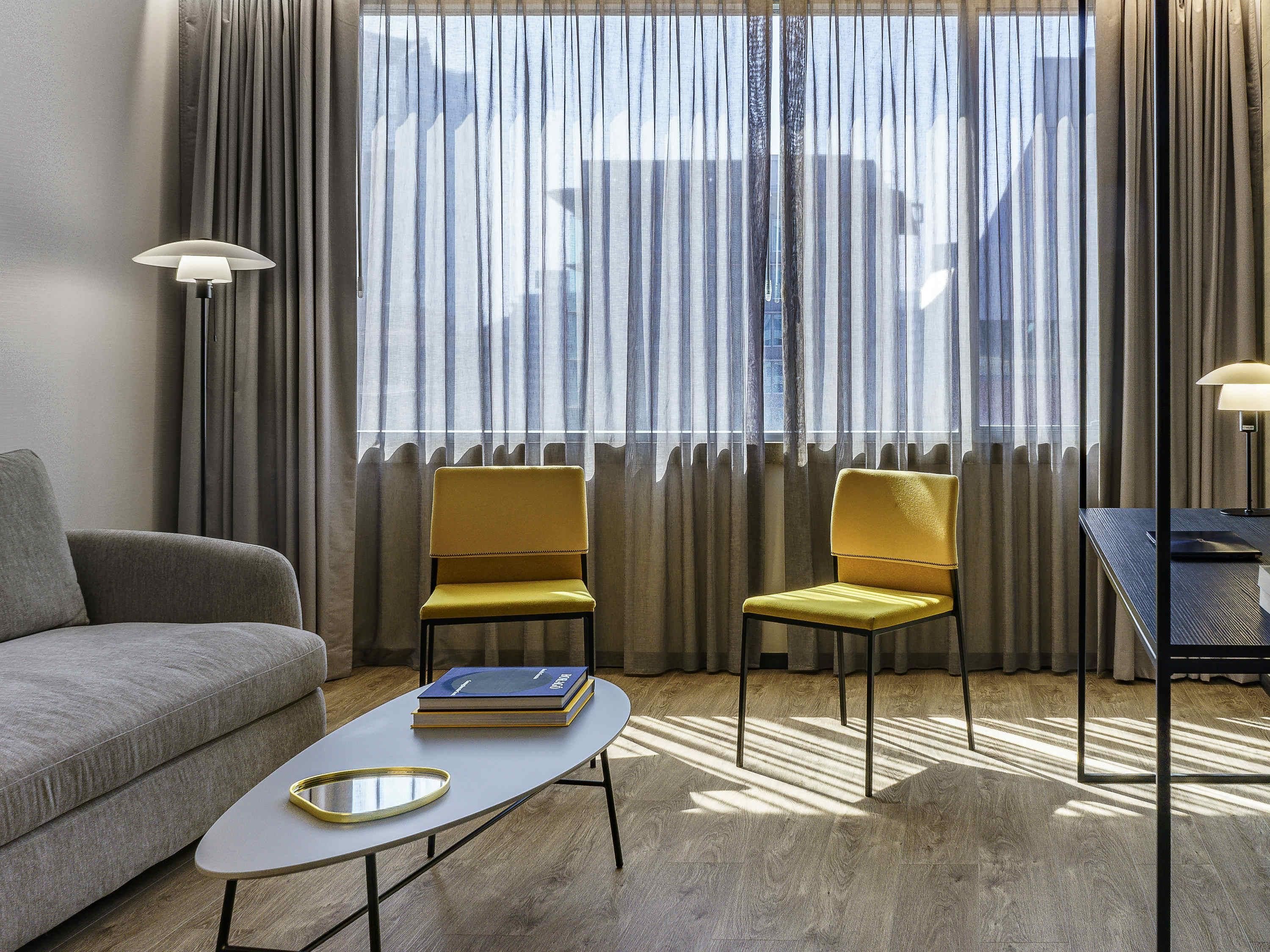 A modern hotel room with a sofa, two yellow chairs, a small table with books, and a desk with a lamp.