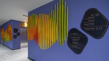 Un pasillo de hotel con diseño moderno, incluyendo una llamativa obra de arte abstracta en la pared y señalización clara para servicios como restaurante, salas de reuniones y habitaciones de huéspedes.