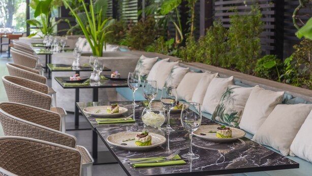 Un restaurante con terraza, mesas de mármol, sillas de mimbre y una fila de bancos acolchados.