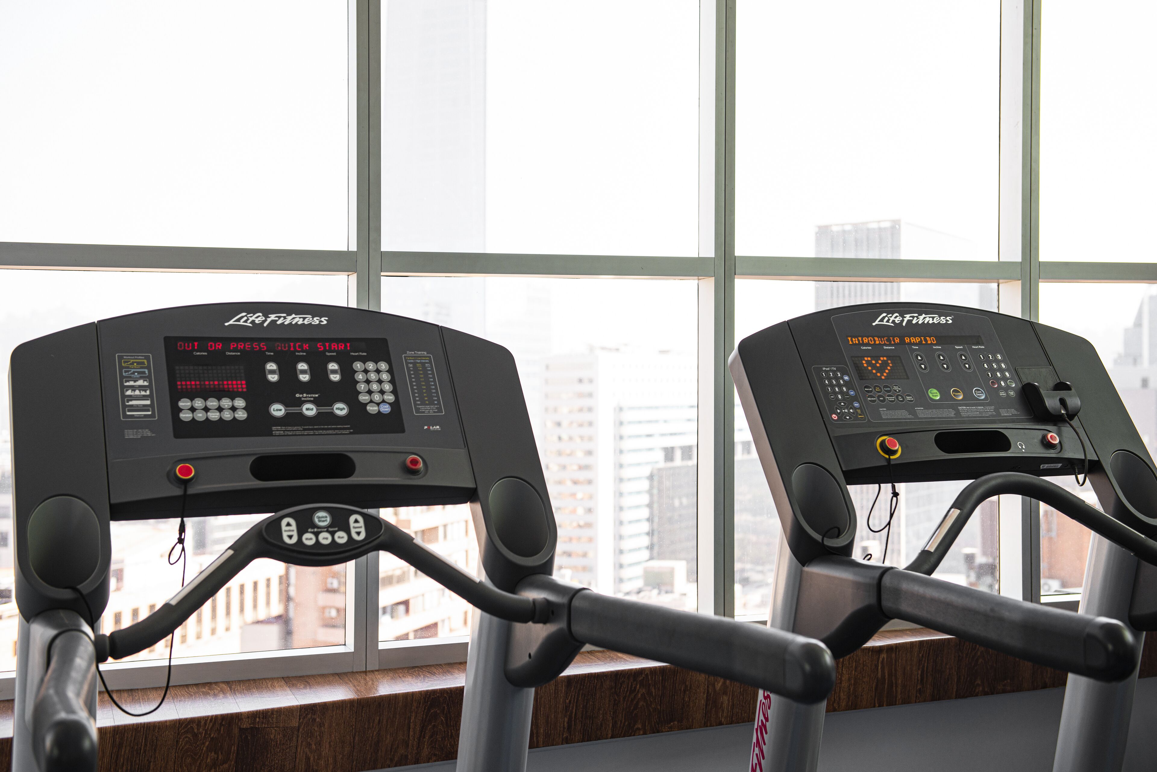 Two Life Fitness treadmills with digital displays, positioned in front of a large window with a city view.