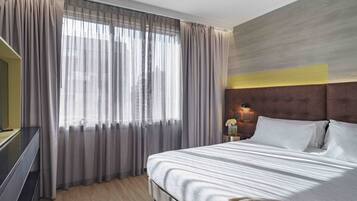A hotel room with a large bed, a TV on a stand, a nightstand with a lamp, and a window with sheer curtains.