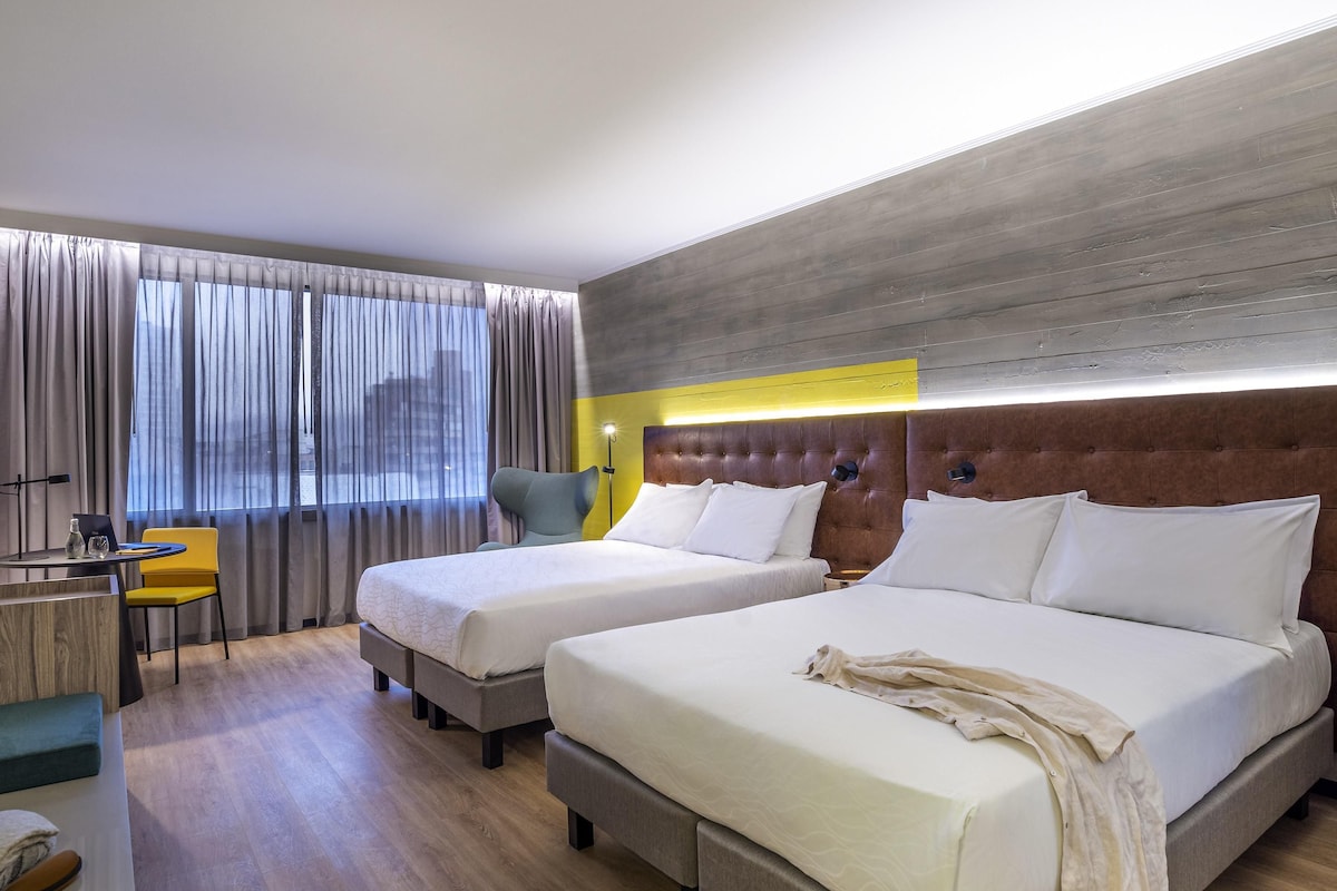 A hotel room with two beds, a desk with a chair, a window with curtains, and a modern headboard.
