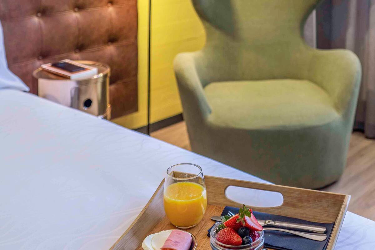 Ein Hotelzimmer mit einem Bett, einem Nachttisch mit Telefon, einem Tablett mit Frühstück, darunter Orangensaft, Erdbeeren und Blaubeeren, sowie einem grünen Sessel.