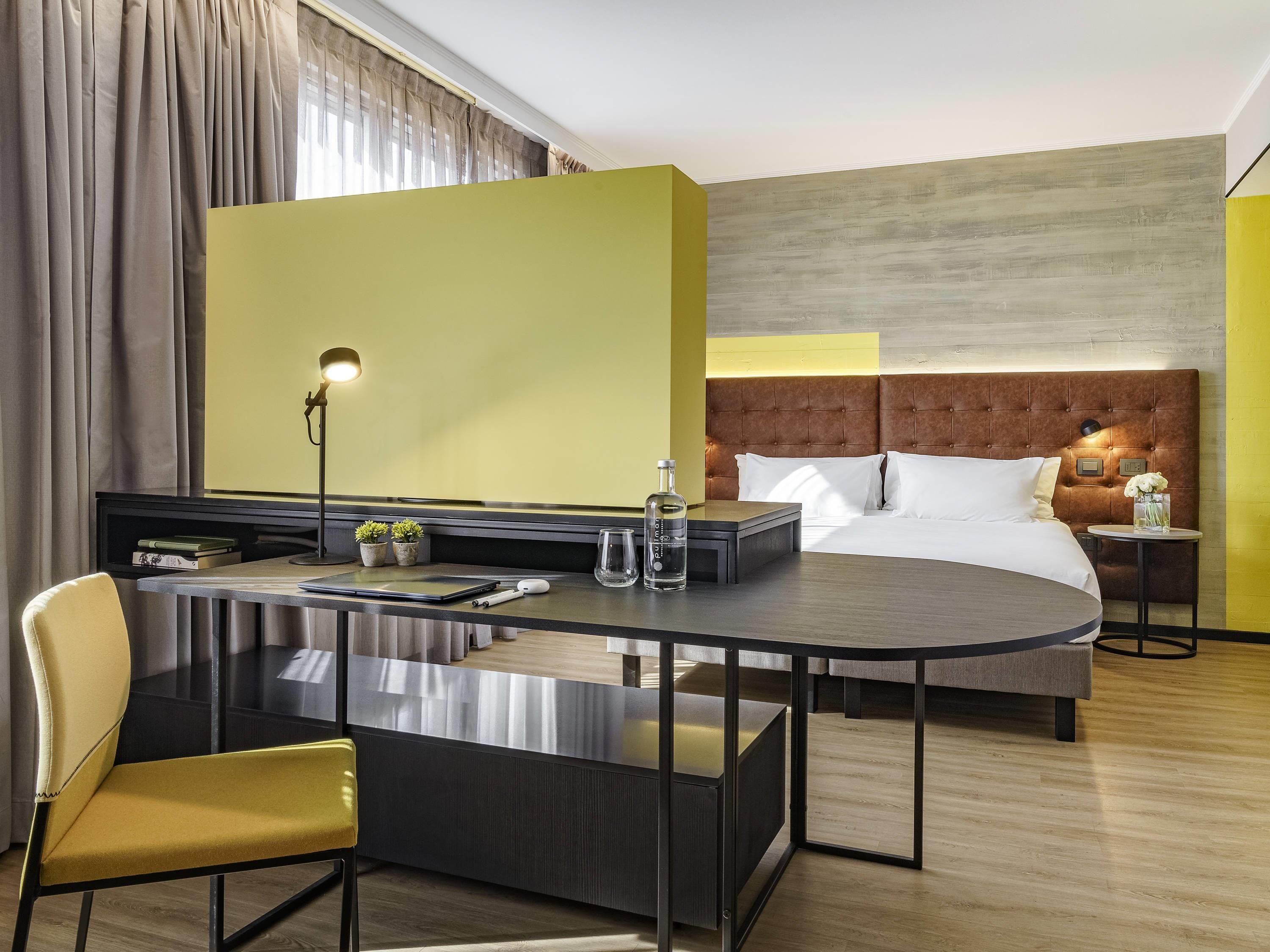 Habitación de hotel con cama, escritorio y silla, lámpara, mesita con un florero y mesita de noche con una flor.